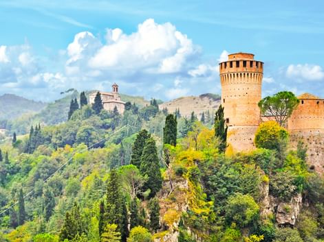 Mittelalterlicher Torre dell'Orologio-Turm auf felsigem Hügel, umgeben von Zypressen und üppiger Vegetation, mit italienischen Hügeln im Hintergrund.