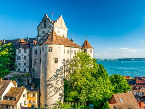 Historische Burg Meersburg mit weißen Mauern und Türmen über rotgedeckten Häusern mit Blick auf das blaue Wasser des Bodensees.