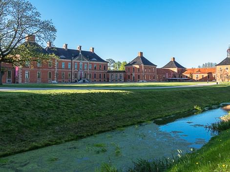 Schloss Klütz mit roten Backsteingebäuden und Wassergraben im Vordergrund. Grüne Rasenfläche und Bäume unter blauem Himmel nahe der Ostsee.
