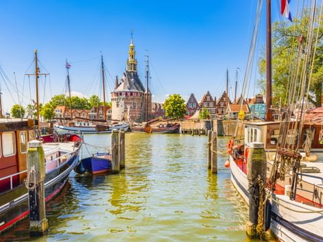Hafenansicht in Hoorn mit traditionellen Booten an Holzpfählen. Historischer Turm und Giebelhäuser säumen das Ufer unter blauem Himmel.