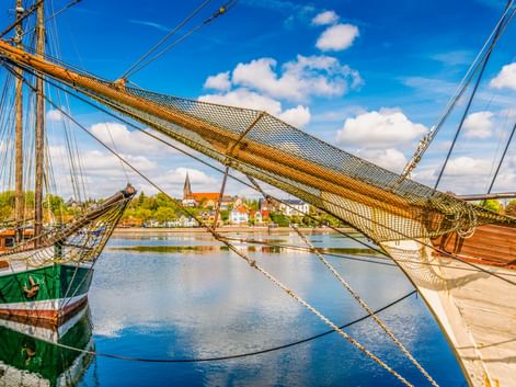 Blick durch die Takelage eines historischen Segelschiffs im Eckernförder Hafen. Das ruhige Wasser spiegelt den blauen Himmel und die Stadt mit Kirchturm.