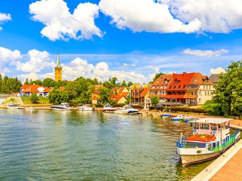 Malerisches Ufer von Mikolajki mit Booten am Ufer, bunten Gebäuden mit roten Dächern und einem Kirchturm unter blauem Himmel.