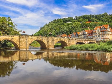 Steinbogenbrücke über die Werra in Hann. Münden mit Fachwerkhäusern und bewaldeten Hügeln, die sich im ruhigen Wasser spiegeln.