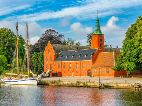 Orangefarbenes Schloss Halmstad mit Uhrturm am Wasser. Ein traditionelles Segelschiff liegt am Kai unter blauem Himmel mit weißen Wolken.
