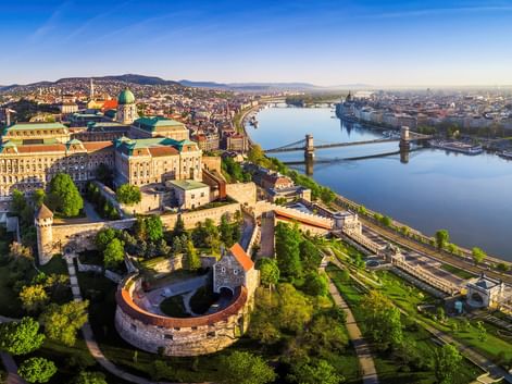 Luftaufnahme von Budapest mit der Donau und Brücken, historischen Gebäuden und dem Parlament in der Ferne unter klarem Himmel.