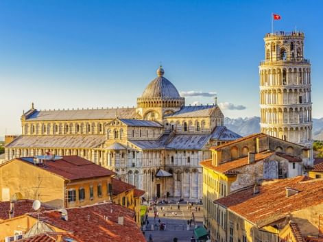 Luftaufnahme des Doms von Pisa mit seiner Kuppel und dem Schiefen Turm über Terrakotta-Dächern unter blauem Himmel.