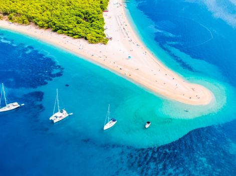 Luftaufnahme vom Strand Zlatni Rat in Bol mit weißer Sandzunge im türkisfarbenen Adriatischen Meer, Segelbooten und grünem Wald.