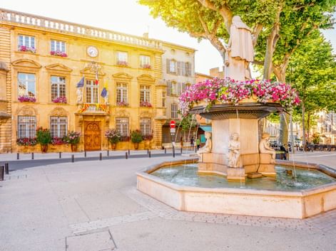 Steinbrunnen mit rosa Blumen in Salon-de-Provence. Gelbes historisches Gebäude mit Uhr und Flaggen im Hintergrund unter Bäumen.
