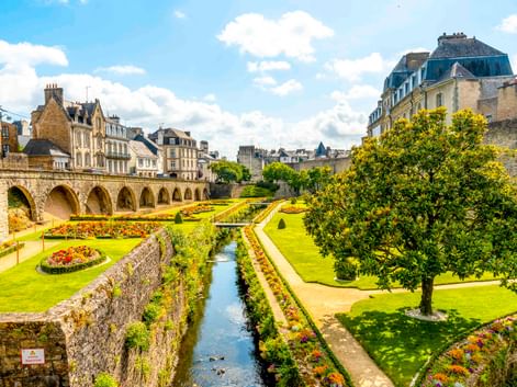 Formale Gärten mit bunten Blumenbeeten und Wasserkanal entlang mittelalterlicher Steinmauern und Bogenbrücke in Vannes, Frankreich.