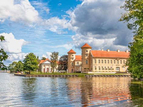Historisches Schloss mit zwei Türmen und rotem Ziegeldach am Seeufer, eingerahmt von grünen Bäumen unter blauem Himmel mit weißen Wolken.