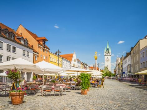 Kopfsteinpflasterplatz in Straubing mit Straßencafés, bunten historischen Gebäuden und dem Stadtturm mit grüner Spitze unter blauem Himmel.