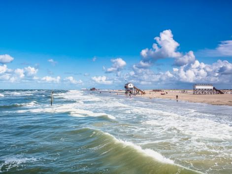 Breiter Sandstrand in St. Peter-Ording mit rollenden Wellen, Pfahlbauten in der Ferne und dramatischen Wolken am blauen Himmel.