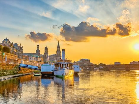 Sonnenuntergang an der Brühlschen Terrasse in Dresden mit Booten an der Elbe. Historische Gebäude und Kirchtürme vor goldenem Himmel.