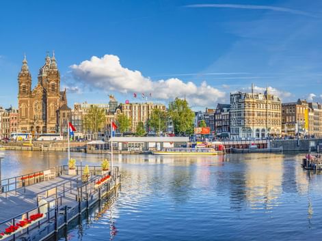 Blick auf Amsterdamer Hafen mit den Zwillingstürmen der St. Nikolaus-Basilika, historischen Gebäuden, Anlegestellen und Booten auf ruhigem Wasser.