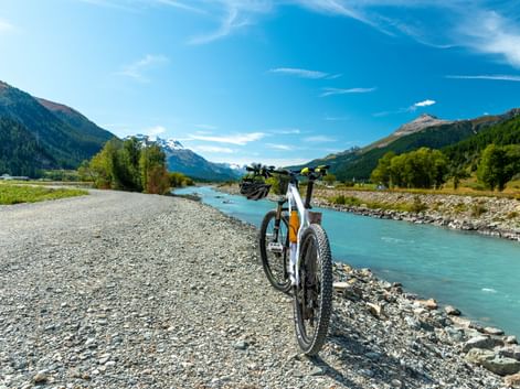 Weißes Fahrrad auf Schotterweg neben türkisfarbenem Fluss bei La Punt. Berge und Wälder rahmen das Tal unter blauem Himmel.