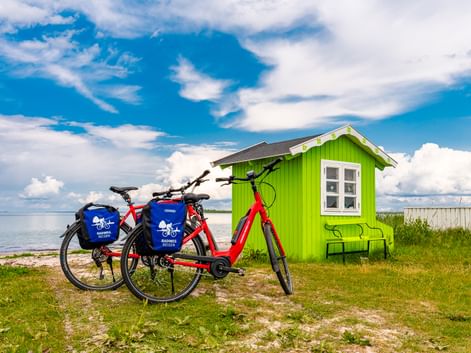 Zwei rote Fahrräder mit blauen Taschen vor einem hellgrünen Strandhaus auf Ærø. Im Hintergrund sind das Meer und der blaue Himmel mit Wolken sichtbar.