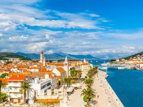 Luftaufnahme der Altstadt von Trogir mit orangefarbenen Dächern, Uferpromenade mit Palmen und hohem Glockenturm. Blauer Hafen und Berge im Hintergrund.