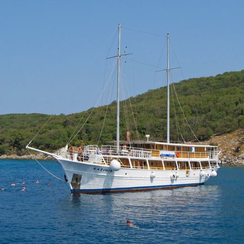 Weißes Motorsegelschiff MS Kazimir mit zwei Masten vor Anker in ruhigem blauen Wasser. Grüne Hügel im Hintergrund, Schwimmer im Wasser.