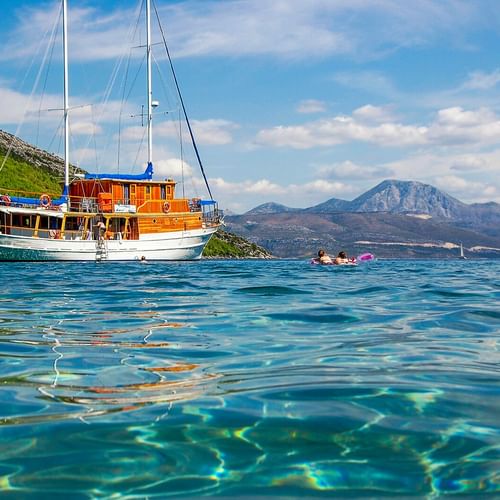 MS Mirabela, ein hölzernes Segelschiff mit weißen Masten, ankert in kristallklarem türkisfarbenem Wasser. Berge und grüne Hügel im Hintergrund.