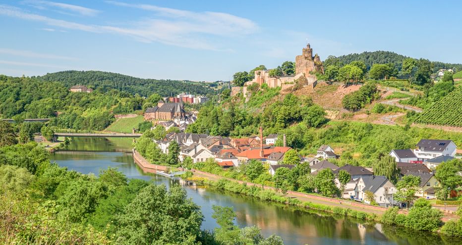 Panoramablick auf Saarburg mit Burgruine auf dem Hügel, Häusern entlang des Flusses, Weinbergen und grünen bewaldeten Hügeln unter blauem Himmel.