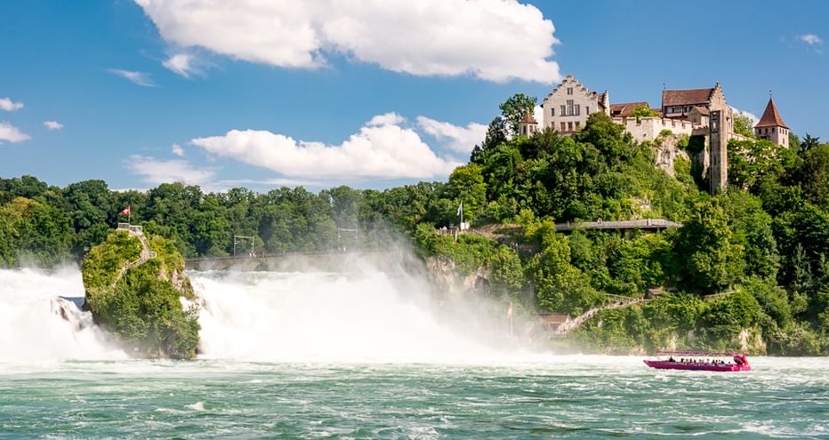 Rheinfall mit massiver Gischt und Schloss Laufen auf grünem Hügel. Ein rotes Touristenboot fährt im türkisfarbenen Wasser unterhalb des Falls.