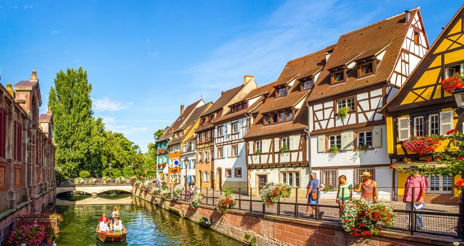 Bunte Fachwerkhäuser entlang eines Kanals in Colmar, Frankreich. Fußgänger spazieren auf der Uferpromenade, während ein kleines Boot auf dem Wasser treibt.
