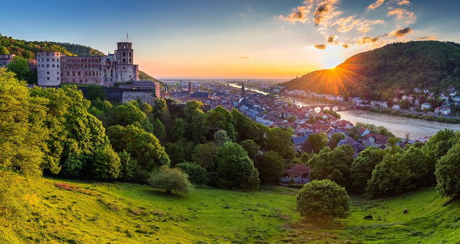 Heidelberger Schloss auf einem Hügel bei Sonnenuntergang mit Blick auf das Neckartal, grünen Wiesen im Vordergrund und historischer Stadt.