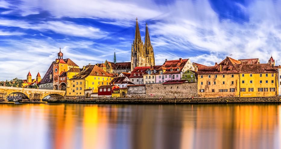 Historische Regensburger Skyline in Bayern mit Domtürmen und bunten Gebäuden, die sich in der Donau unter dramatischem Himmel spiegeln.