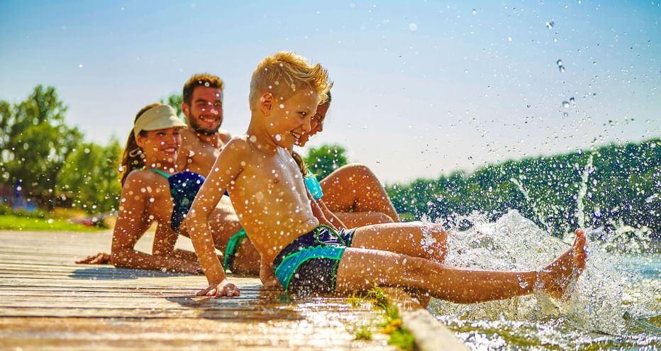 Familie mit zwei Kindern planscht auf einem Holzsteg am Brombachsee, mit klarem blauen Himmel und grünen Bäumen im Hintergrund.