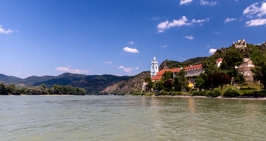 Blick auf die Donau mit dem blau-weißen Turm von Stift Dürnstein und Burgruine auf einem Hügel. Berge und blauer Himmel im Hintergrund.