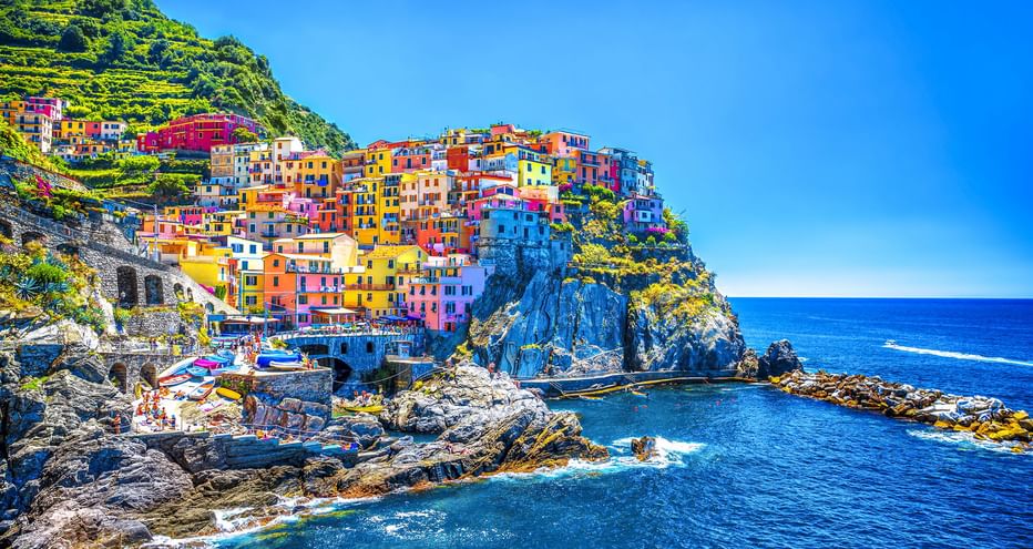 Leuchtend bunte Häuser eines Cinque Terre Dorfes auf felsigen Klippen über dem blauen Mittelmeer unter klarem Himmel.