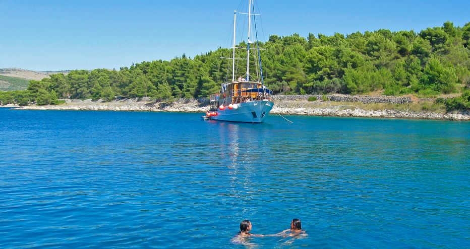 MS Mirabela, ein blaues Segelschiff mit zwei Masten, ankert in ruhigen kroatischen Gewässern. Zwei Schwimmer im Vordergrund, grüne Küste dahinter.