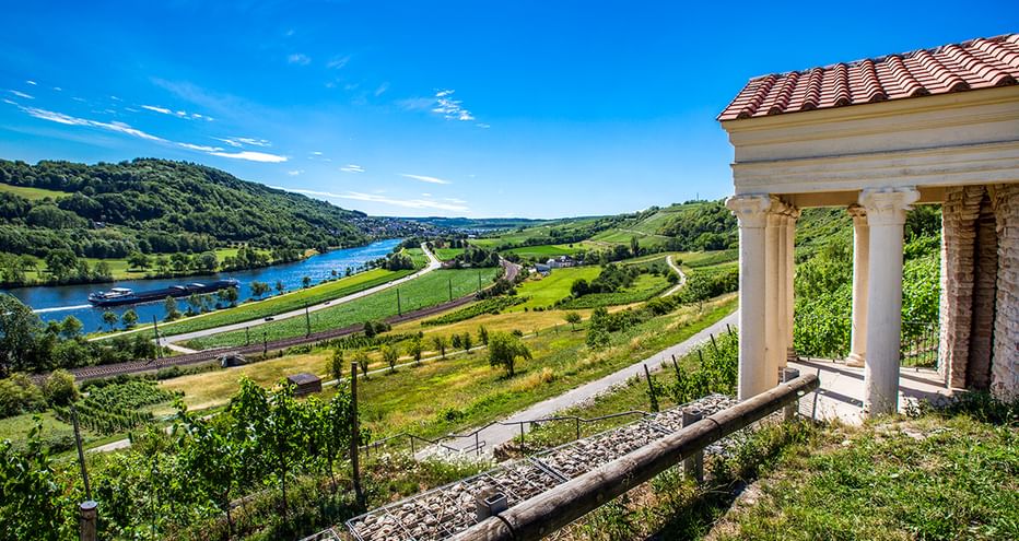 Malerischer Blick auf die Mosel, die sich durch grüne Hügel und Weinberge schlängelt, mit einem Pavillon mit Ziegeldach und weißen Säulen im Vordergrund.