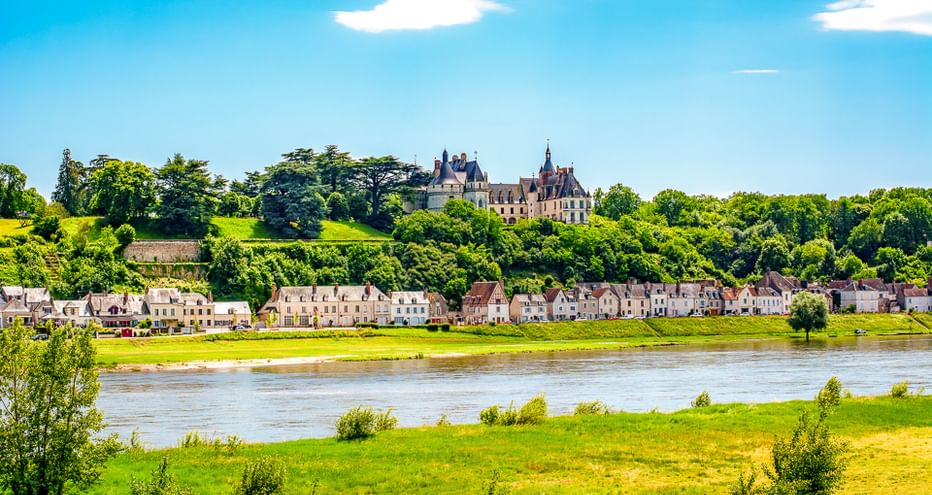Französisches Schloss auf Hügel über Dorfhäusern entlang der Loire. Grüne Wiesen im Vordergrund unter blauem Himmel mit weißen Wolken.