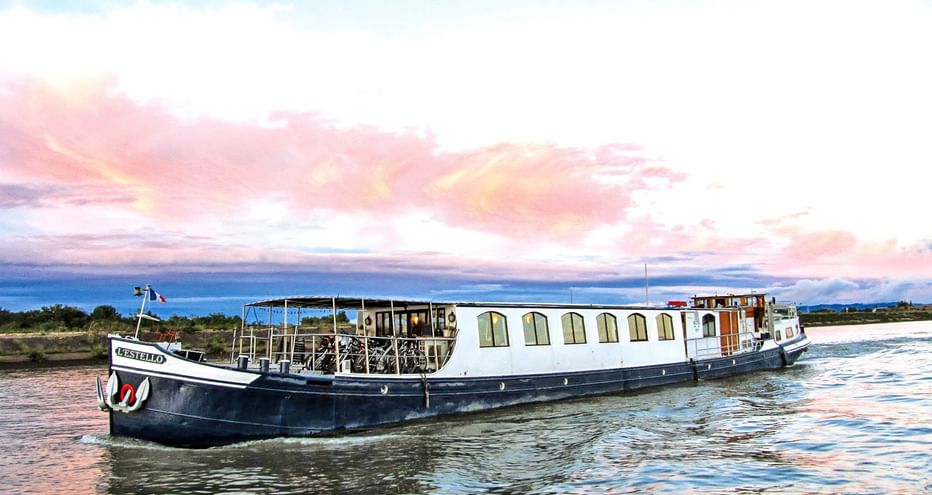 Weiß-blaues Flussschiff L'Estello mit Fahrrädern an Deck auf ruhigem Wasser in der Provence unter rosa-blauem Abendhimmel.