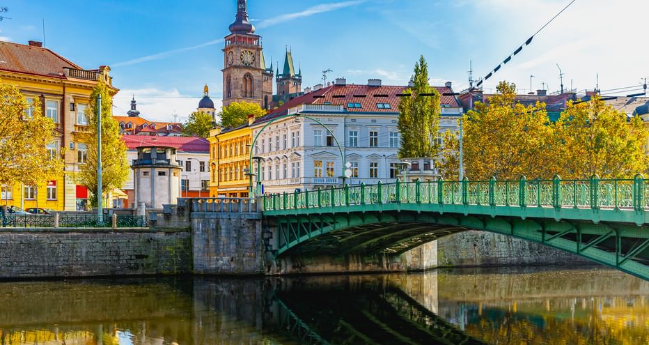 Grüne Zierbrücke über die Elbe mit bunten historischen Gebäuden und Kirchturm in einer tschechischen Stadt unter blauem Himmel.