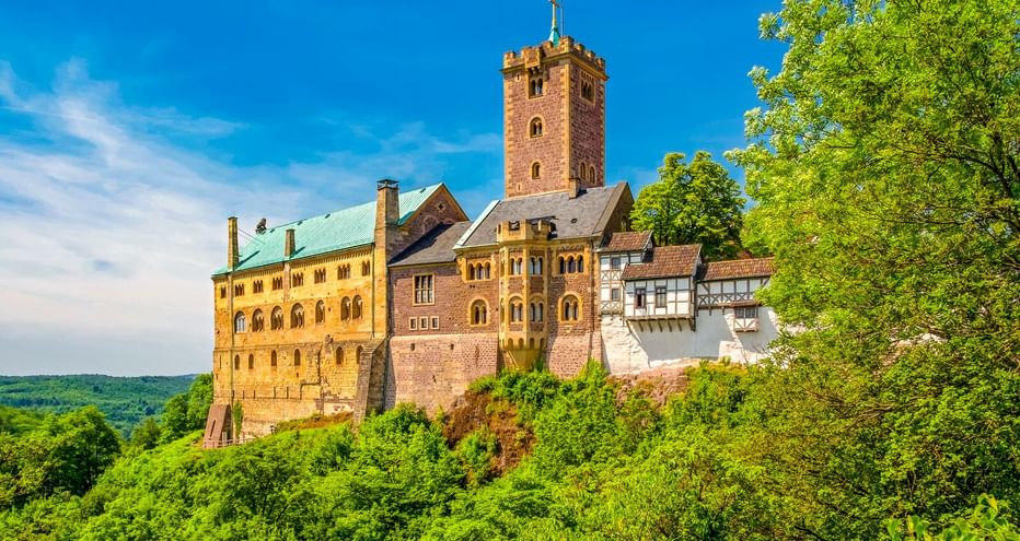 Die Wartburg in Eisenach thront auf einem bewaldeten Hügel mit rotem Turm, Steingebäuden und Fachwerkhäusern unter blauem Himmel.