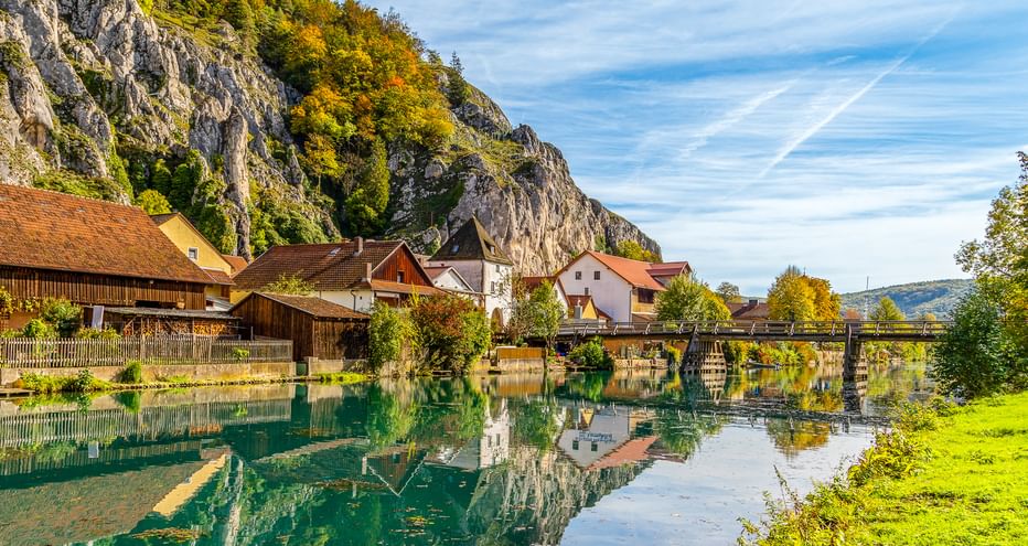 Dorf Essing am Altmühl-Fluss mit traditionellen Häusern, Holzbrücke und dramatischen Kalksteinfelsen in Herbstfarben.