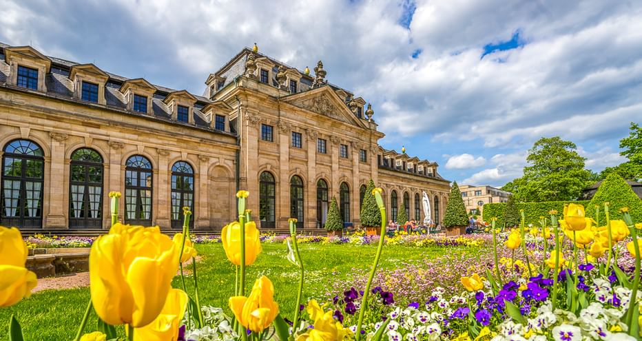 Barockes Palais mit verzierter Fassade und blühendem Garten mit gelben Tulpen und lila Blumen unter bewölktem Himmel.