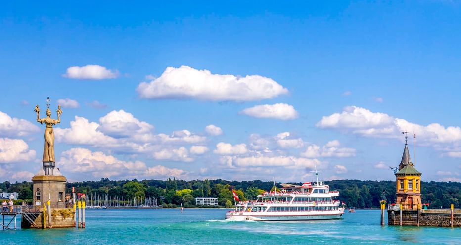 Weiße Personenfähre fährt durch die Konstanzer Hafeneinfahrt zwischen zwei Türmen. Links: goldene Imperia-Statue, rechts: Leuchtturm mit gelbem Turm.