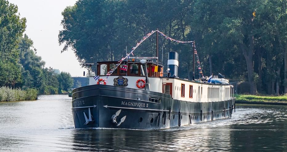 Passagierschiff Magnifique II mit dunklem Rumpf und weißer Kabine auf ruhigem Kanal, gesäumt von dichten grünen Bäumen unter klarem Himmel.