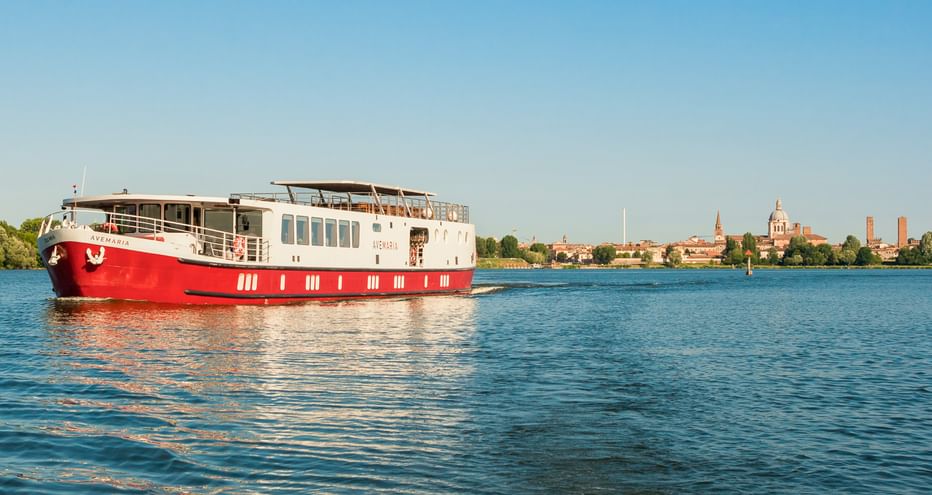 Rot-weißes Flusskreuzfahrtschiff MS Ave Maria auf ruhigem blauen Wasser mit Mantuas historischer Skyline im Hintergrund unter klarem Himmel.