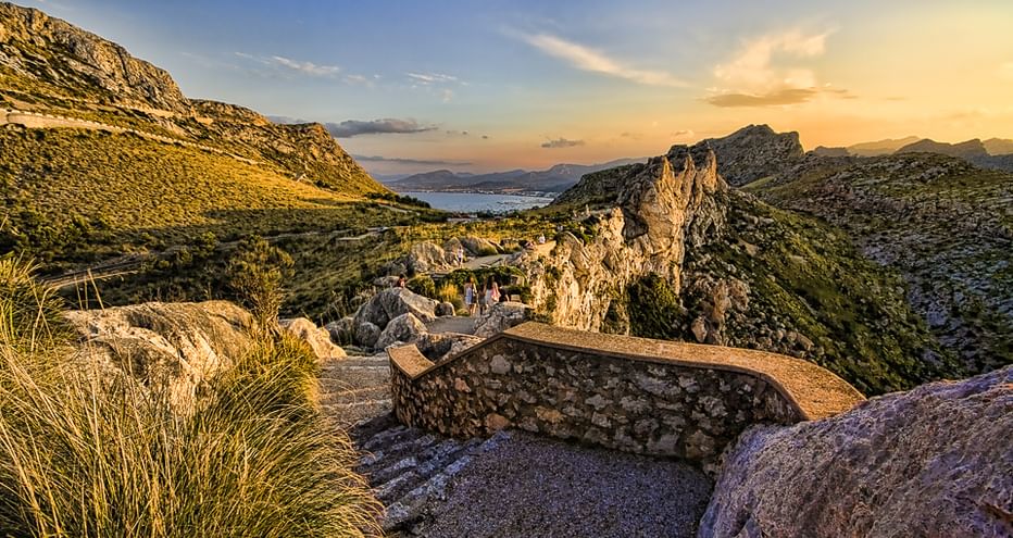 Steinerner Aussichtspunkt mit Felsformationen in Mallorcas Bergen bei Sonnenuntergang, mit Blick auf Küstenlandschaft und Meer unter goldenem Himmel.