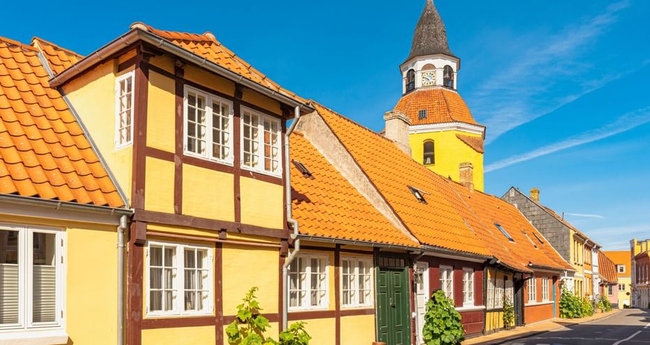 Historische Straße in Faaborg mit gelben Fachwerkhäusern und orangen Ziegeldächern. Kirchenglockenturm mit Uhr im Hintergrund unter blauem Himmel.