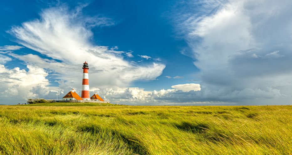 Leuchtturm Westerhever mit rot-weißen Streifen und angrenzenden Gebäuden in grünen Salzwiesen unter dramatischem blauen Himmel mit weißen Wolken.