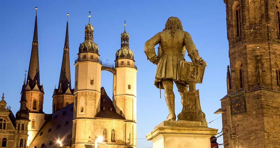 Bronzestatue eines Mannes mit Schwert auf dem Marktplatz in Halle an der Saale, mit beleuchteten Kirchtürmen vor blauem Abendhimmel.