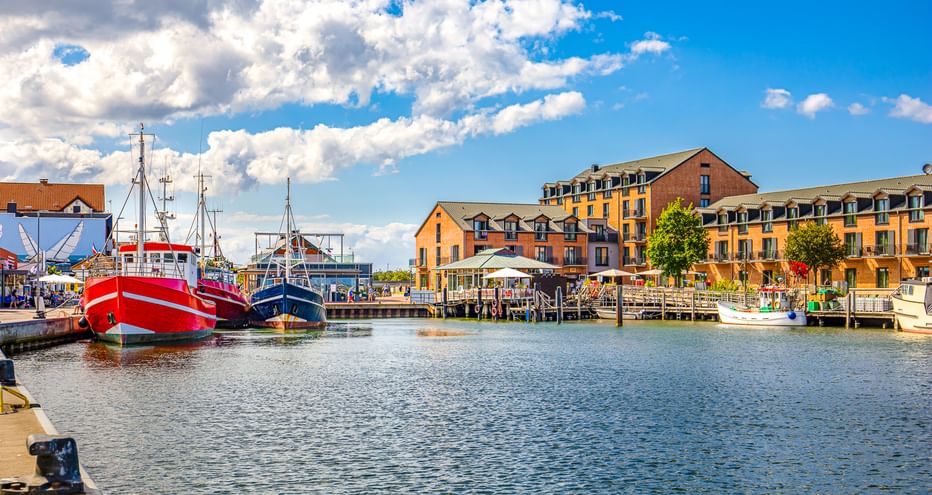 Heiligenhafener Hafen mit roten und blauen Fischerbooten am Kai. Bunte Gebäude säumen die Uferpromenade unter blauem Himmel mit Wolken.