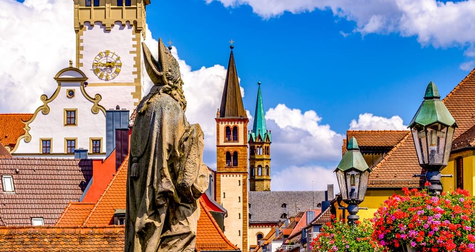 Steinstatue eines Heiligen auf der Alten Mainbrücke mit Würzburger Altstadtsilhouette, Kirchtürmen, Uhrturm und roten Ziegeldächern.