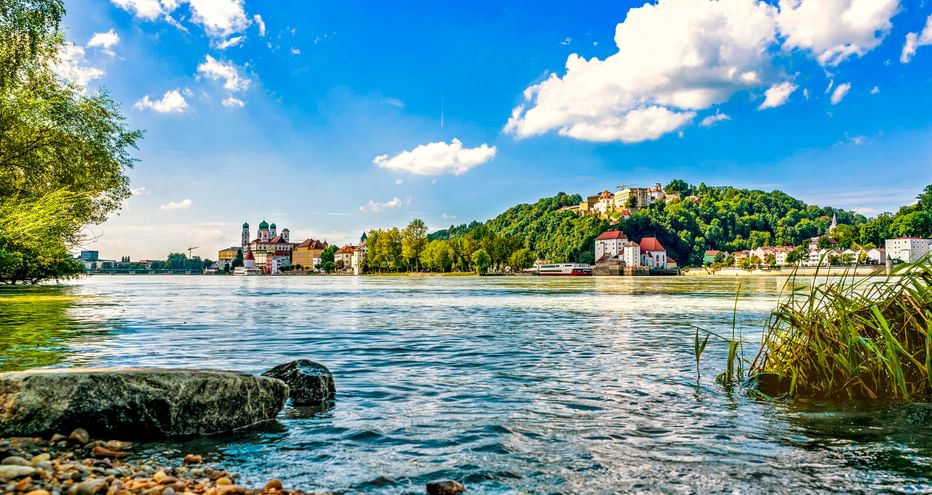 Blick über die Donau in Passau mit Kieselstrand, historischen Gebäuden und Dom links sowie einer Festung auf dem Hügel unter blauem Himmel.
