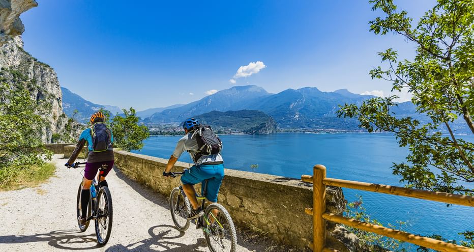 Zwei Radfahrer auf einem Panoramaweg mit Holzgeländer über dem Gardasee. Berge und blaues Wasser im Hintergrund unter klarem Himmel.